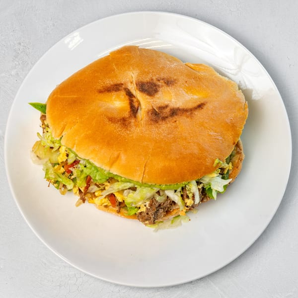 A Muchas Gracias Mexican Food torta, filled with meat, scrambled eggs, lettuce, and avocado, on a white plate.