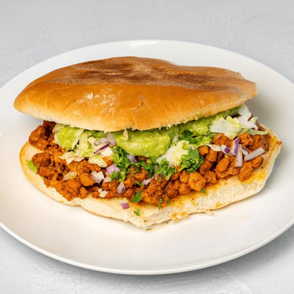 Muchas Gracias Mexican Food's torta with seasoned meat, guacamole, lettuce, and onions on a white plate.