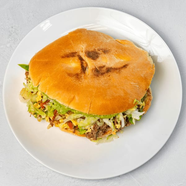A Muchas Gracias Mexican Food torta, filled with meat, lettuce, and avocado, on a white plate.